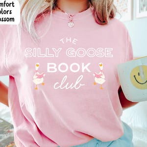 Può includere: Una maglietta rosa chiaro con la scritta bianca "THE SILLY GOOSE BOOK CLUB". La maglietta presenta illustrazioni di oche che leggono libri. Nella foto è tenuta una tazza bianca con una faccina sorridente gialla.