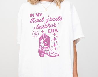Op de afbeelding: Wit T-shirt met roze tekst die "In my third grade teacher era" luidt, met een roze cowboylaars met sterren en een halve maan. De tekst "Yee Haw" staat in een cirkel boven de laars.
