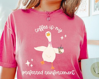 May include: A pink t-shirt with the text "coffee is my preferred reinforcement" and a cartoon goose holding a coffee cup. The shirt also has the word "CRUNCH BERRY" printed at the bottom.