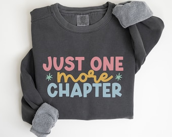 May include: A dark gray sweatshirt with the text "JUST ONE *more* CHAPTER" in colorful letters. The sweatshirt has a fleece lining on the inside of the sleeves.