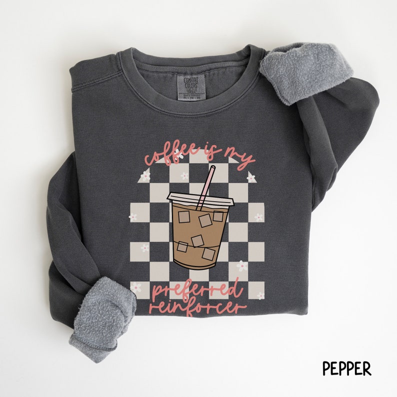 May include: A dark gray sweatshirt with the text "coffee is my preferred reinforcer" and a graphic of an iced coffee drink. The design includes a checkered background and floral accents. The sweatshirt is labeled "pepper".