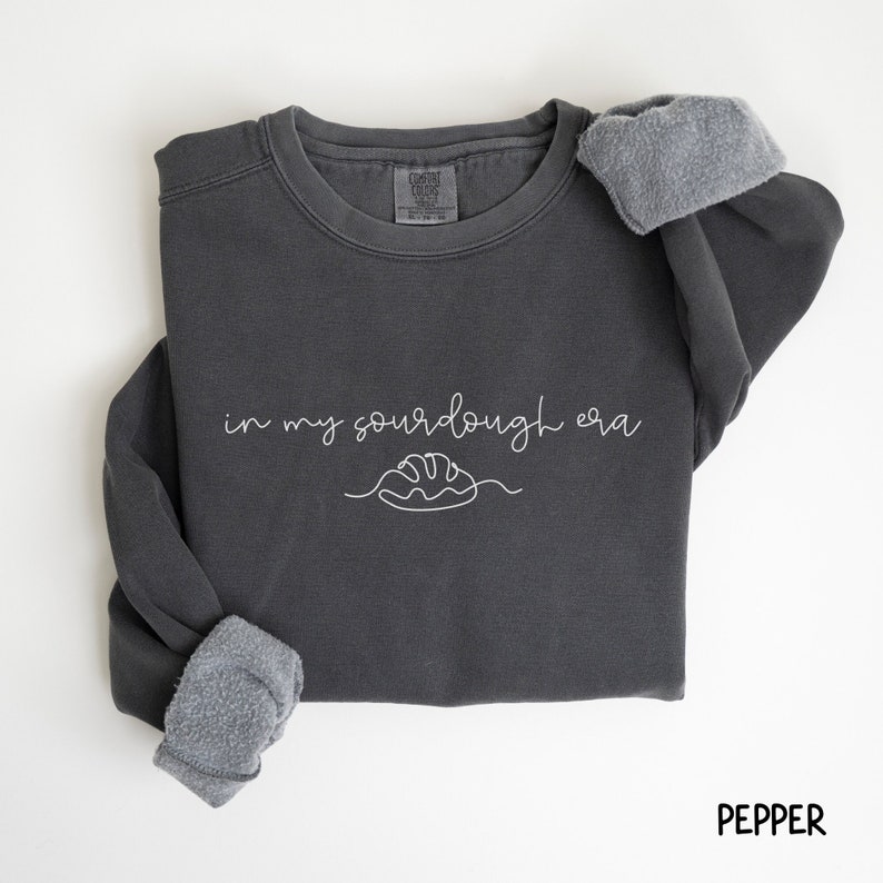Peut inclure: Un sweat-shirt gris fonc&eacute; avec le texte "in my sourdough era" et un dessin simple d'une miche de pain. Le sweat-shirt est fait d'un tissu doux et confortable et a une coupe d&eacute;contract&eacute;e.