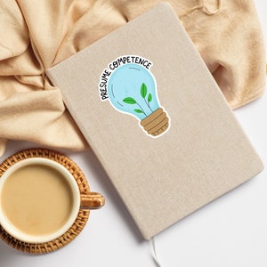 Op de afbeelding: Een lichtbruin notitieboekje met een lichtblauwe sticker in de vorm van een gloeilamp waarop "Presume Competence" staat. De gloeilamp heeft een groene plant die erin groeit.