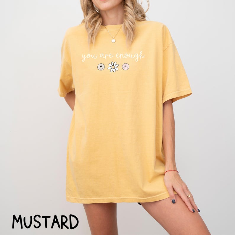 May include: A mustard yellow t-shirt with the text "you are enough" in white lettering. The shirt features a small white daisy with three smiley faces on either side.