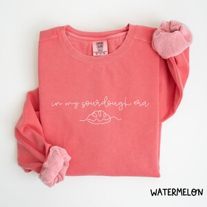Peut inclure: Un sweat-shirt rose corail avec le texte "in my sourdough era" en &eacute;criture blanche avec un dessin simple d'une miche de pain.