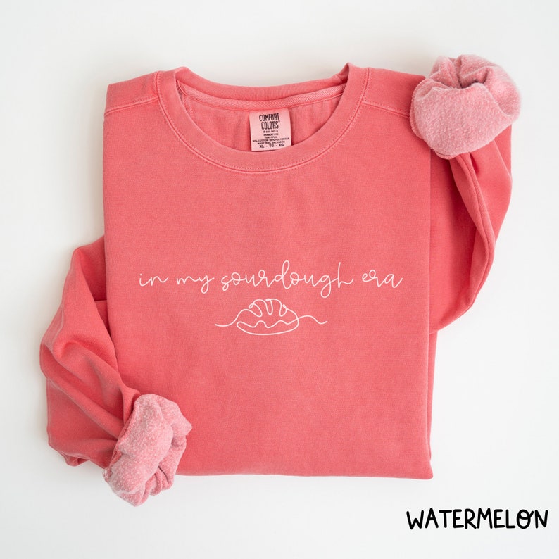Peut inclure: Un sweat-shirt rose corail avec le texte "in my sourdough era" en &eacute;criture blanche avec un dessin simple d'une miche de pain.
