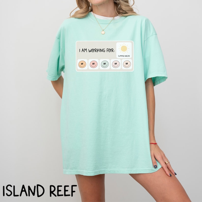 May include: A light blue t-shirt with a graphic print that says "I am working for: Summer Break". The graphic features a sun and a row of six smiley faces, each with a different color.