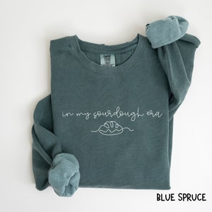 Peut inclure: Un sweat-shirt bleu sarcelle avec le texte "in my sourdough era" et un dessin simple d'une miche de pain. Le sweat-shirt est pli&eacute; et les manches sont retrouss&eacute;es.