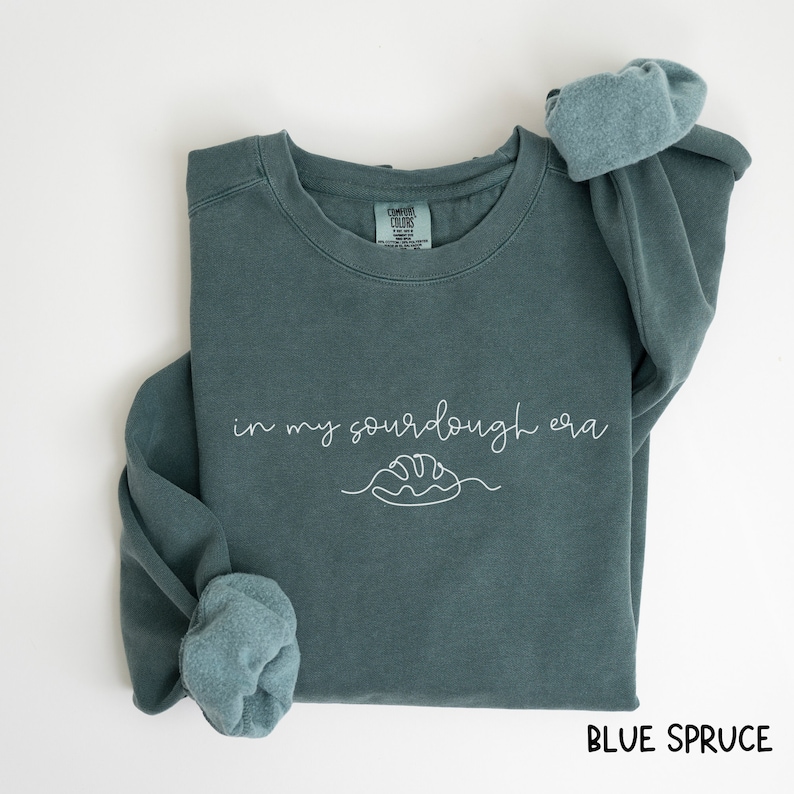 Peut inclure: Un sweat-shirt bleu sarcelle avec le texte "in my sourdough era" et un dessin simple d'une miche de pain. Le sweat-shirt est pli&eacute; et les manches sont retrouss&eacute;es.