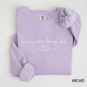 Peut inclure: Un sweat-shirt lavande avec le texte "in my sourdough era" et un dessin simple d'une miche de pain.