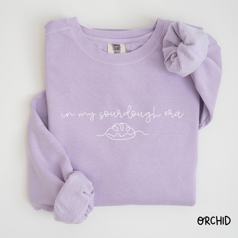 Peut inclure: Un sweat-shirt lavande avec le texte "in my sourdough era" et un dessin simple d'une miche de pain.