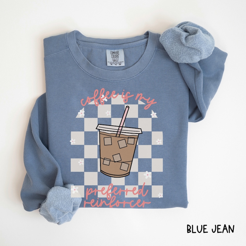 May include: Blue jean-colored sweatshirt with the text "coffee is my preferred reinforcer" in pink script. The design features a coffee cup with ice cubes on a checkered background. A scrunchie rests on the sleeve.