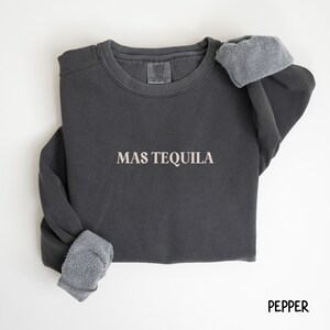 May include: A dark gray sweatshirt with the text "MAS TEQUILA" in white lettering. The sweatshirt has a fleece lining and is made of a soft, comfortable material.