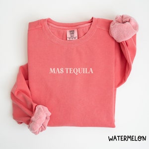May include: A pink sweatshirt with the text "MAS TEQUILA" in white lettering. The sweatshirt is made of a soft, comfortable fabric and has a relaxed fit.