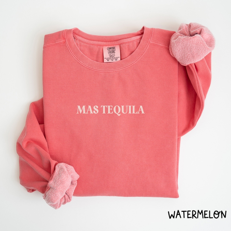 May include: A pink sweatshirt with the text "MAS TEQUILA" in white lettering. The sweatshirt is made of a soft, comfortable fabric and has a relaxed fit.