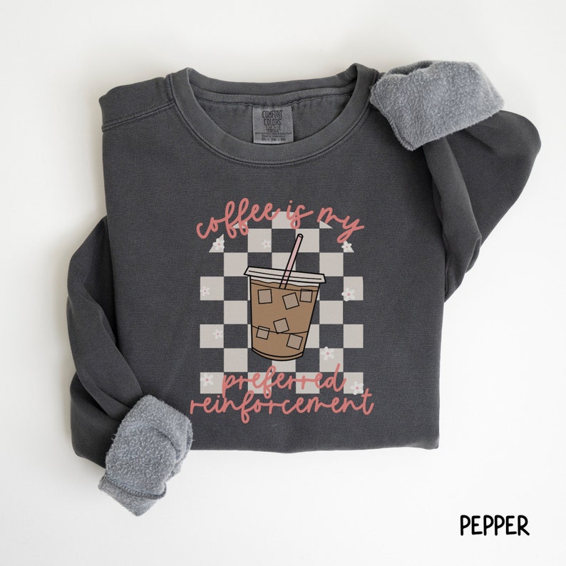 Puede incluir: Una sudadera gris oscuro con el texto "coffee is my preferred reinforcement" en rosa. El texto est&aacute; rodeado de un patr&oacute;n a cuadros y una imagen de dibujos animados de una taza de caf&eacute; helado.