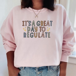 Peut inclure: Un sweat-shirt rose avec le texte "It's a great day to regulate" en lettres color&eacute;es avec des &eacute;toiles de chaque c&ocirc;t&eacute; du mot "day".