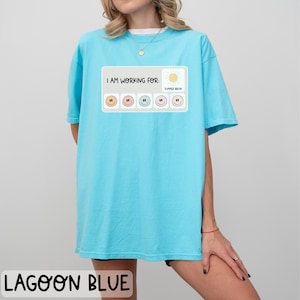 May include: A light blue t-shirt with a graphic that says "I am working for: Summer Break" with a series of smiley faces in different colors.
