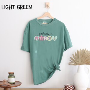 May include: Light green t-shirt with a graphic of the word "pediatric nurse" in colorful letters with a star and heart design.