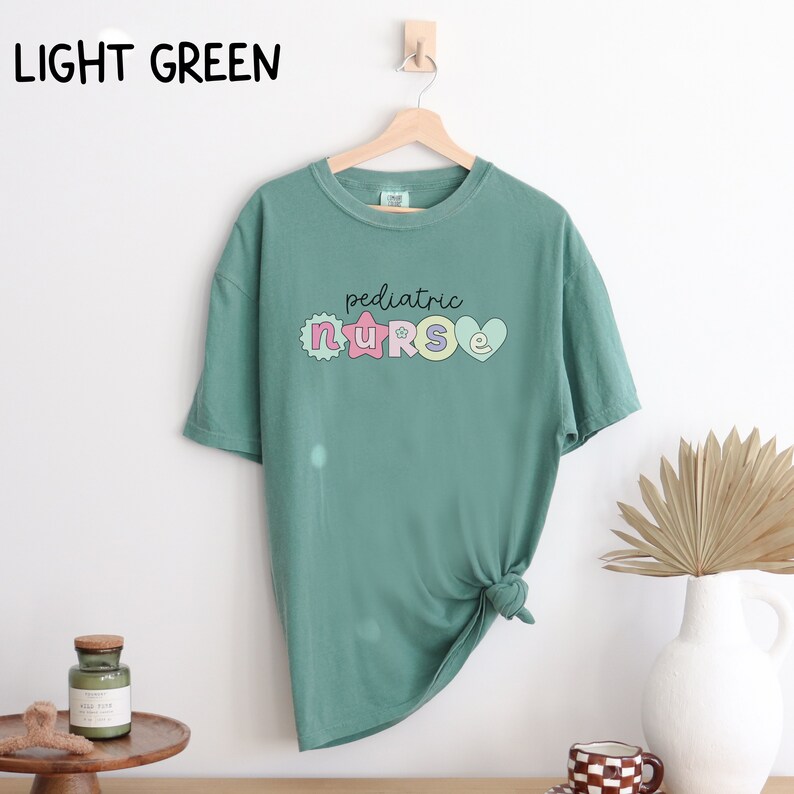 May include: Light green t-shirt with a graphic of the word "pediatric nurse" in colorful letters with a star and heart design.