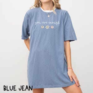 May include: A blue jean t-shirt with the text "you are enough" in white lettering. The text is accompanied by three small graphic icons: a smiley face, a daisy, and a heart.