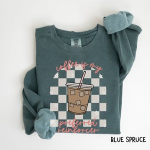 May include: Blue spruce sweatshirt with the text "coffee is my preferred reinforcer" and a graphic of an iced coffee drink. The design includes a checkered background and small white flowers. The sweatshirt is a Comfort Colors brand.