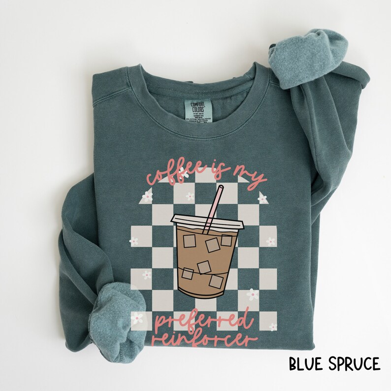 May include: Blue spruce sweatshirt with the text "coffee is my preferred reinforcer" and a graphic of an iced coffee drink. The design includes a checkered background and small white flowers. The sweatshirt is a Comfort Colors brand.