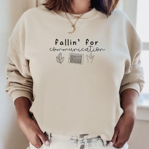 Può includere: Una felpa beige con la scritta "fallin' for communication" stampata sul davanti. Il testo è accompagnato da tre icone: una foglia, un tablet e una mano che fa il segno "Ti amo".