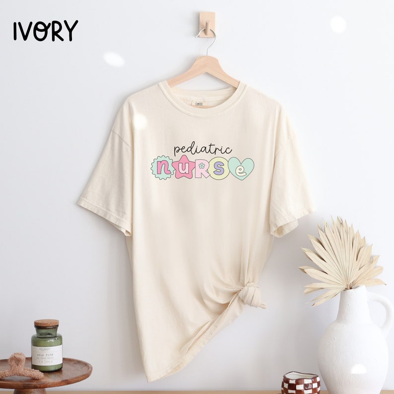 May include: A cream-colored t-shirt with the text "pediatric nurse" in black lettering. The letters are decorated with pastel colors and designs, including a star, a flower, and a heart.