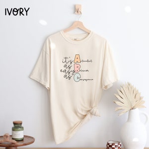 Op de afbeelding: Een crèmekleurig T-shirt met korte mouwen met de tekst "it's as easy as ABC" met de letters A, B en C in verschillende kleuren en de woorden "antecedent", "gedrag" en "gevolg" naast elke letter.