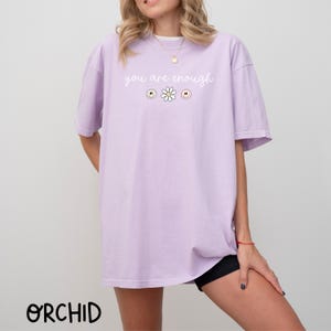 May include: A lavender t-shirt with the text "you are enough" in white lettering. The shirt features a small daisy graphic with a yellow centre and white petals.