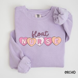 May include: A lavender sweatshirt with the text "float nurse" in pink and orange letters inside heart shapes.