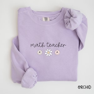 Könnte beinhalten: Ein lavendelfarbener Pullover mit dem Text "math teacher" und drei Smiley-Gesichtern, eines mit einem rosa Gesicht, eines mit einem weißen Gesicht und eines mit einem Pfirsich-Gesicht. Eine Gänseblümchen befindet sich zwischen den beiden Smiley-Gesichtern mit rosa Gesichtern.