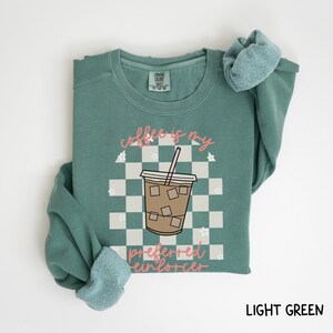 May include: Light green sweatshirt with the text "coffee is my preferred reinforcer" and a graphic of an iced coffee drink. The design is set against a checkered background with floral accents. The sweatshirt is folded.