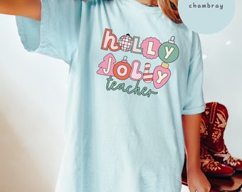 May include: A light blue t-shirt with the text "holly jolly teacher" in colorful, festive lettering. The text is surrounded by Christmas-themed illustrations, including ornaments, a tree, and a disco ball.