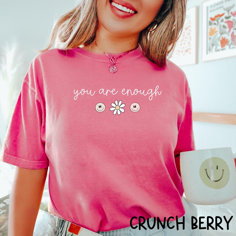 May include: Pink t-shirt with the text "you are enough" in white lettering. The shirt also features three smiley faces and a daisy.