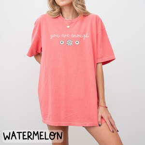 May include: An oversized pink t-shirt with the text "you are enough" in white lettering. The shirt features three smiley faces and a daisy.