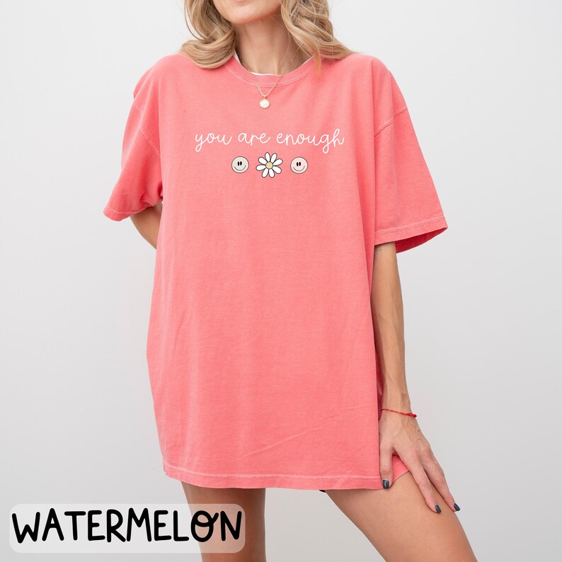 May include: An oversized pink t-shirt with the text "you are enough" in white lettering. The shirt features three smiley faces and a daisy.