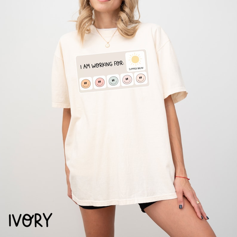 May include: A white t-shirt with the text "I AM WORKING FOR: SUMMER BREAK" and a graphic of a sun and six smiley faces. The shirt is on a model wearing black shorts.