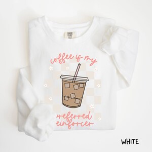 May include: White sweatshirt with the phrase "coffee is my preferred enforcer" in pink script. The design includes a coffee cup graphic with ice cubes and a pink straw, set against a checkered background with small flower accents. The word "WHITE" is at the bottom right.