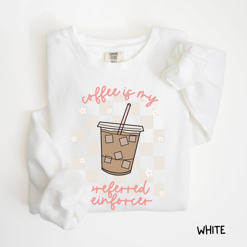 May include: White sweatshirt with the phrase "coffee is my preferred enforcer" in pink script. The design includes a coffee cup graphic with ice cubes and a pink straw, set against a checkered background with small flower accents. The word "WHITE" is at the bottom right.