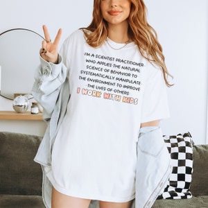 May include: White t-shirt with a pink and orange graphic that reads "I'm a scientist practitioner who applies the natural science of behavior to systematically manipulate the environment to improve the lives of others I work with kids."