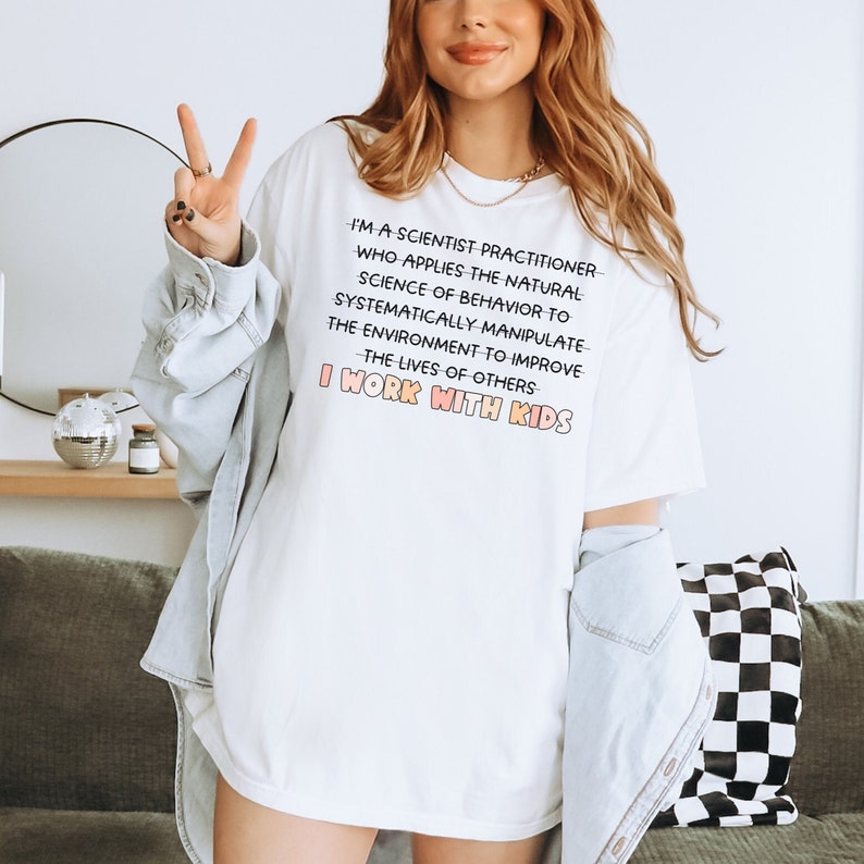 May include: White t-shirt with a pink and orange graphic that reads "I'm a scientist practitioner who applies the natural science of behavior to systematically manipulate the environment to improve the lives of others I work with kids."