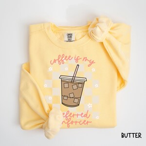 May include: A butter-yellow sweatshirt with the words "coffee is my preferred enforcer" in pink script. The design features a coffee cup with ice cubes on a checkered background, and small white flowers. The word "BUTTER" is printed in the bottom right corner.