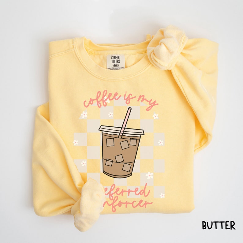May include: A butter-yellow sweatshirt with the words "coffee is my preferred enforcer" in pink script. The design features a coffee cup with ice cubes on a checkered background, and small white flowers. The word "BUTTER" is printed in the bottom right corner.