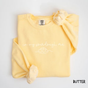 Peut inclure: Un sweat-shirt jaune avec le texte "in my sourdough era" et un dessin simple d'une miche de pain. Le sweat-shirt est pli&eacute; et l'&eacute;tiquette indique "Comfort Colors".