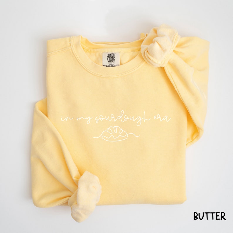 Peut inclure: Un sweat-shirt jaune avec le texte "in my sourdough era" et un dessin simple d'une miche de pain. Le sweat-shirt est pli&eacute; et l'&eacute;tiquette indique "Comfort Colors".