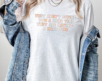 Puede incluir: Una camiseta gris de manga larga con el texto "They aren't giving you a hard time they are having a hard time" impreso en letras de colores.