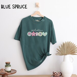 May include: A teal green t-shirt with a graphic design that says "pediatric nurse" in colorful letters. The letters are decorated with hearts, stars, and flowers.