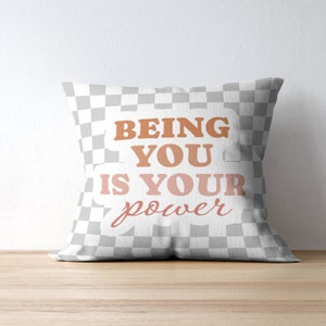 Può includere: Un cuscino bianco con un motivo a scacchi e il testo "BEING YOU IS YOUR power" in un gradiente rosa e arancione.